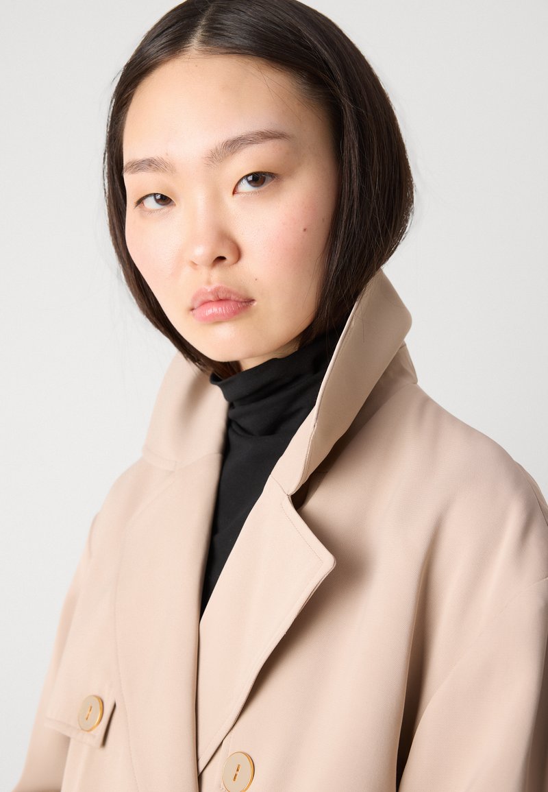 Beige Trenchcoat mit einem geschlitzten Kragen, doppelter Knopfleiste und hellbraunen Knöpfen, über einem schwarzen Rollkragenpullover getragen. Glatte Stoffoberfläche.