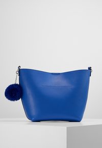 Borsa in pelle blu con una silhouette curva, caratterizzata da una catenina e un pom pom blu. Texture liscia e design minimalista senza hardware visibile.