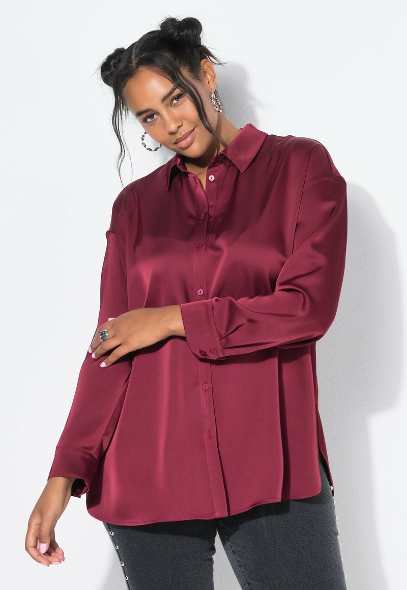 Camisa de satén burdeos con botones, de manga larga, con cuello clásico y aberturas laterales, combinada con jeans oscuros con detalles de botones.