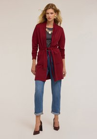 Burgundy cardigan med knytband i midjan, ribbade detaljer och långa ärmar, parat med ljusblå jeans med uppvikta ben och burgundy högklackade skor.