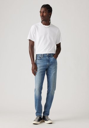 Tricou alb din bumbac, asortat cu blugi din denim bleu deschis. Blugii au o croială dreaptă și sunt purtați cu adidași alb-negru.