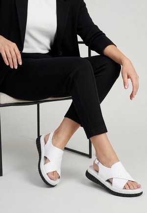 Person iført sorte bukser og jakke, hvid top, siddende på stol med benene krydset, iført hvide sandaler med sorte såler.