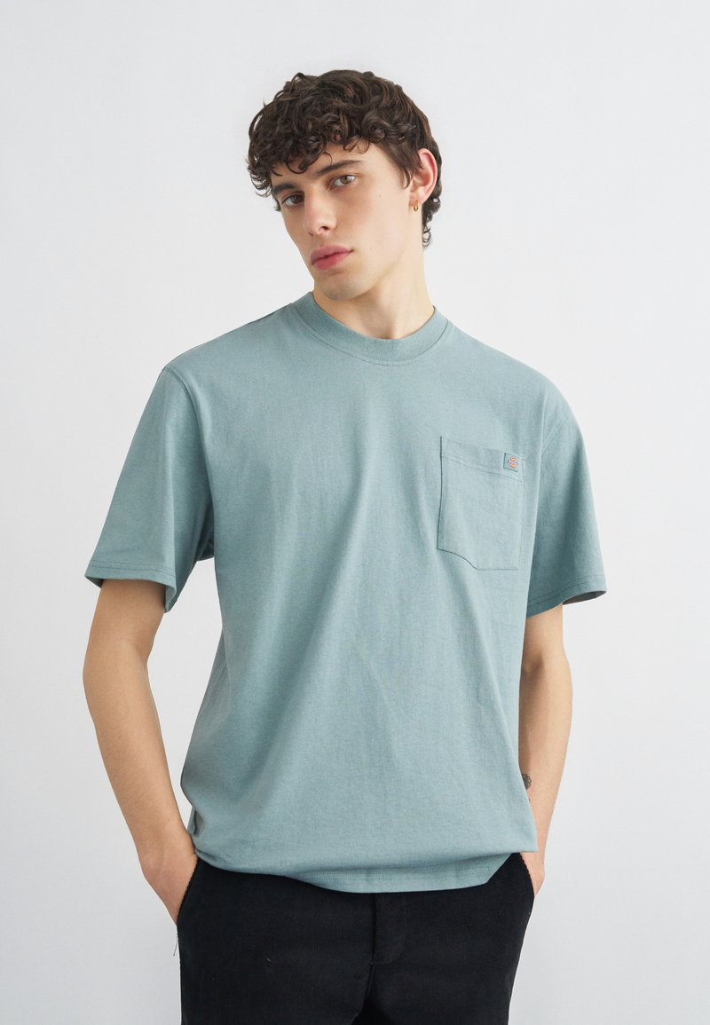 Giovane uomo con capelli ricci che indossa una maglietta a maniche corte azzurro chiaro con una tasca sul petto, mani nelle tasche dei pantaloni neri, sfondo neutro.