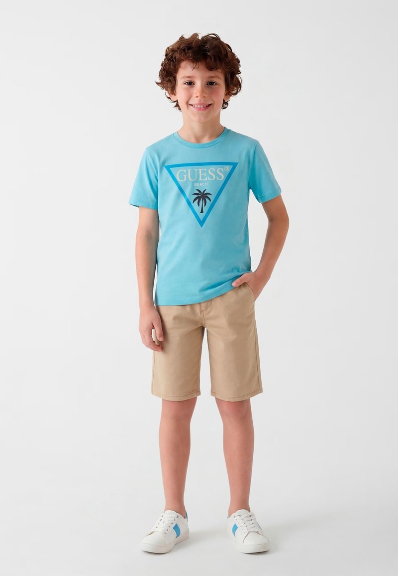 Garçon aux cheveux bouclés debout en souriant, portant un t-shirt bleu avec le logo "Guess Beach", un short beige et des baskets blanches sur un fond uni.