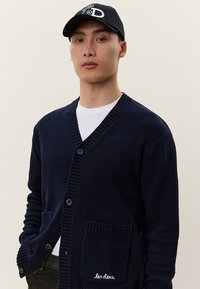 Άνδρας που φοράει μπλε σκούρο ζακέτα με κουμπιά και τσέ�πες, λευκό πουκάμισο, σκουρόχρωμα τζιν και μαύρο καπέλο με λευκές γράμματα.