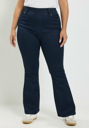 Personne portant un jean bootcut bleu foncé, des baskets blanches et un haut blanc, debout devant un fond clair uni.