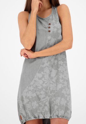 Kvinde iført grå ærmeløs kjole med tre knapper nær halsudskæringen og subtilt blomsterprint på den nederste halvdel, stående med armene over kors.