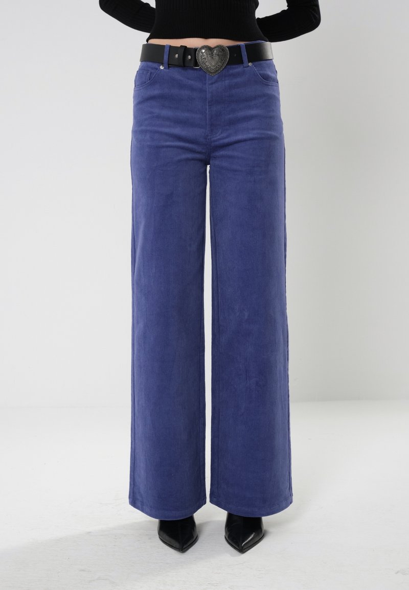 Vidbeniga, högmidjade lila kordbyxor med en slät yta, prydda med ett svart bälte med en hjärtformad spänne.