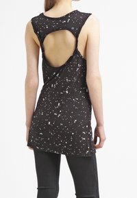 Svart ärmlös topp med vitt sprutmönster, med en utskärning på ryggen. Mjuk tyg och avslappnad passform med förlängd nederkant.