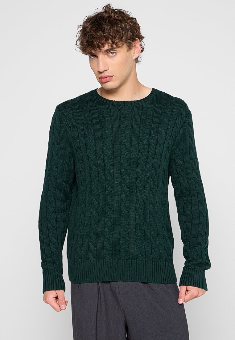 Suéter de punto cable en un verde oscuro, con cuello redondo, mangas largas y puños y dobladillo acanalados. Presenta un pequeño acento de logo.