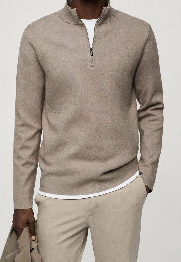 Homme portant un pull beige à demi-fermeture éclair par-dessus une chemise blanche, accompagné d'un pantalon beige assorti, tenant une veste beige dans sa main gauche.