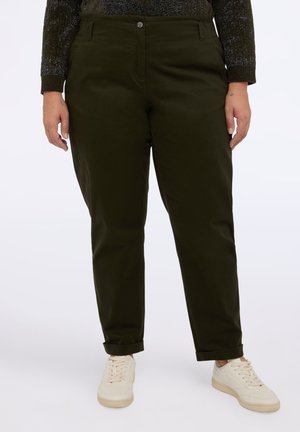 Persona con pantaloni dritti color oliva scuro arrotolati alle caviglie e sneaker bianche, in piedi contro uno sfondo semplice.