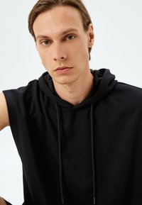 Sudadera sin mangas negra hecha de tela suave, con capucha ajustable y textura lisa, de diseño minimalista y sin patrones visibles.