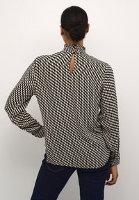Langearmige blouse in een zwart en crèmekleurig houndstooth patroon, met een hoge halslijn met plooidetails en een sleutelgatopening aan de achterkant.