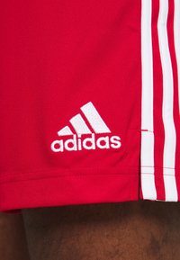 Short de sport rouge avec logo Adidas blanc et trois bandes blanches verticales sur le côté, porté sur une jambe.