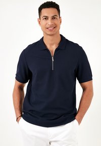 Cămașă polo cu mânecă scurtă, de culoare bleumarin, cu guler cu fermoar, realizată dintr-un material texturat; prezintă o croială modernă și un design minimalist.