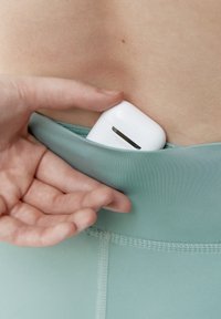 Trådlös öronsnäcka i ett vitt etui, som vilar i en ficka av ljus turkos tyg. En hand rör försiktigt vid kanten av fickan, vilket visar hur väl den passar.