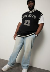 Schwarze Strickweste mit "THE MYTH" und "21" in Grau, getragen über einem weißen Hemd. Hellblaue Denim-Hosen und weiße Schuhe vervollständigen das Outfit.