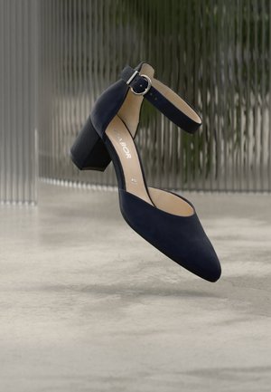 Zapato de mujer de ante negro con tira en el tobillo y tacón cuadrado, flotando sobre un suelo liso de hormigón con un fondo de cristal texturizado.
