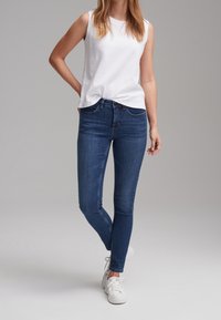Femme portant un haut blanc sans manches, un jean slim bleu et des baskets blanches, debout, jambes croisées et une main dans le dos.