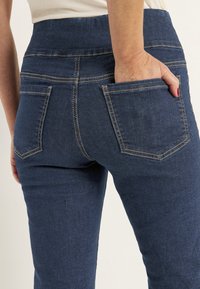 Mörkblå denimjeans med hög midja, synlig sömnad och bakfickor. Texturen är slät, designad för en åtsittande silhuett.