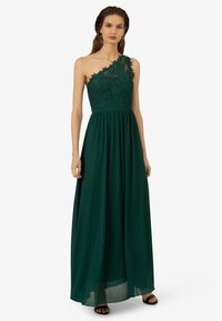 Ein einseitig tragendes, smaragdgrünes Kleid mit Spitzenverzierungen am Oberteil, fließendem Rock und taillierter Fit, kombiniert mit schwarzen Sandalen.