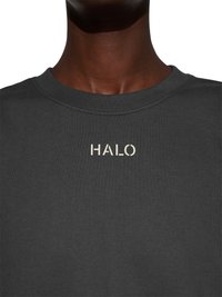 Mørk hudfarvet person iført en mørkegrå crewneck-trøje med ordet "HALO" trykt i lyse bogstaver på brystet.