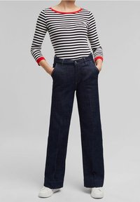 Gestreiftes langärmeliges Oberteil mit rotem Saum, kombiniert mit hochtaillierten, dunkelblauen, weiten Denim-Hosen. Das Model trägt weiße Sneaker.