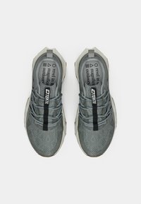 Graue Sportschuhe mit gemustertem Mesh-Obermaterial, reflektierenden Akzenten, einem schwarzen Markenriemen und einer strukturierten Sohle, von oben betrachtet.