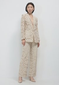 Donna che indossa un completo in pizzo floreale beige con giacca e pantaloni a gamba larga, abbinato a tacchi alti beige, in piedi contro uno sfondo bianco.