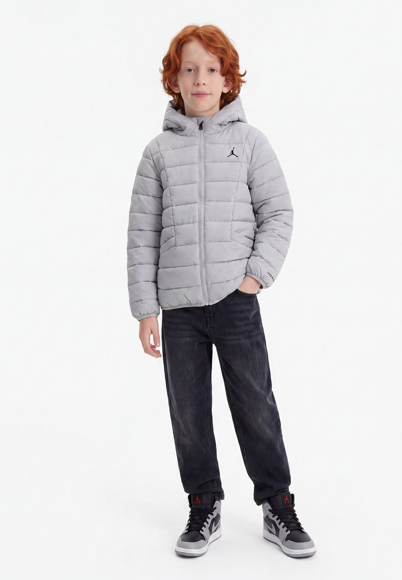 Enfant aux cheveux roux portant une doudoune grise à capuche, un jean noir et des baskets montantes grises et noires, debout avec une main dans la poche.