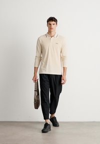 Mann som går iført beige langermet polo, svarte cropped bukser, svarte sneakers, og bærer en brun skulderveske foran en enkel hvit vegg.