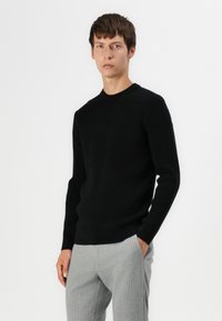 Suéter negro de punto con un cuello redondo, mangas largas y puños acanalados, combinado con pantalones gris a rayas.