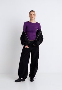 Μωβ μπλούζα με μακριά μανίκια συνδυασμένη με μαύρο, φαρδύ bomber jacket και χαλαρό μαύρο cargo παντελόνι. Μαύρα παπούτσια ολοκληρώνουν το ντύσιμο.