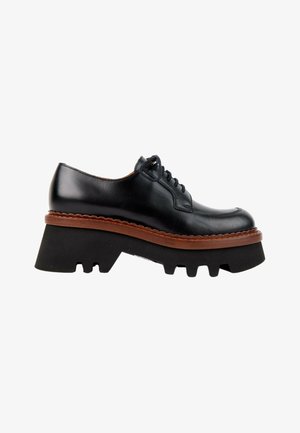 Chaussure noire en cuir à lacets avec une semelle plateforme noire épaisse et une bordure brunâtre en forme d'ondulations sur la semelle intercalaire, vue de profil.