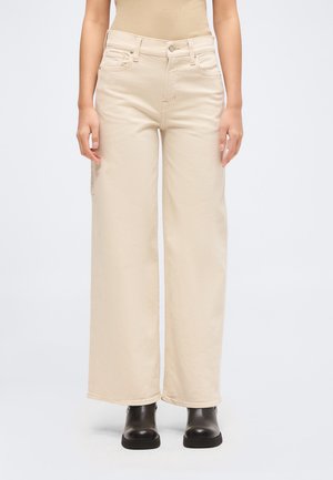 Žena nosí krémové nohavice s vysokým pásom a širokými nohavicami v kombinácii s čiernymi kotníkovými topánkami, stojí na jednolivom svetlom pozadí.