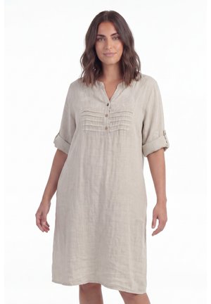 Femme portant une robe en lin beige longueur genou, manches retroussées, boutons et détails brodés sur la poitrine, debout sur fond blanc.