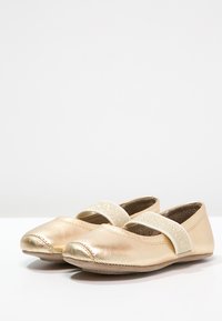 Gylne lærballetsko med en glitrende elastisk stropp. Har en rund tå og sydd detaljer på sålen. Glatt tekstur.