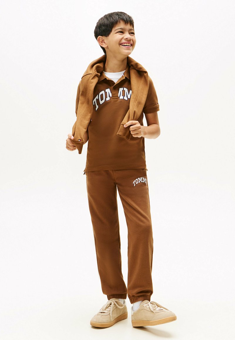 Ensemble de sport en coton marron comprenant un polo avec logo et un pantalon de jogging. La tenue a une coupe décontractée et est complétée par des chaussures beiges.