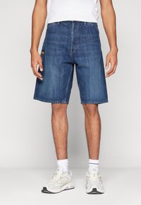 Hombre con pantalones cortos de mezclilla azul de media longitud, calcetines blancos hasta el tobillo, zapatillas deportivas blancas y una camiseta blanca metida por dentro, todo frente a un fondo liso.