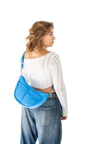 Borsa a spalla in nylon blu con forma semi-circolare, dotata di tasca con zip e tracolla regolabile, indossata sopra una maglietta bianca testurizzata e jeans.