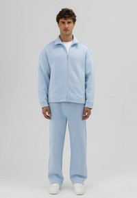 Veste zippée bleu clair avec col montant et manches larges, assortie à un pantalon ample en tissu doux. Baskets blanches.