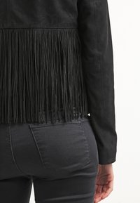 Veste en daim noir avec détails à franges à l'arrière, manches longues et silhouette ajustée. Associée à un jean foncé avec une poche arrière.