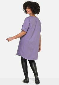 Paarse tuniek tot de knie met korte opgerolde mouwen, gecombineerd met glanzende zwarte leggings en gestifte enkellaarzen. Glad textuur; comfortabele pasvorm.
