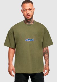 Olīvu zaļš oversized T-krekls, izgatavots no kokvilnas, ar krāsainu, stilizētu grafisko logo centrā. Īsas piedurknes un apaļš kakls.