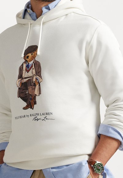 Homem com um hoodie branco com um gráfico do Polo Bear, vestido com um conjunto vintage, camisa azul claro por baixo e um relógio verde com bracelete de couro castanho.