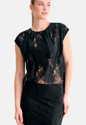 Femme portant un haut en dentelle noire à manches courtes et une jupe assortie en dentelle noire, regardant légèrement sur le côté.