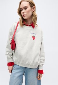 Νεαρή γυναίκα που φοράει ανοιχτό γκρι φούτερ με μια κόκκινη φράουλα και κείμενο, κόκκινο πουκάμισο από κάτω, ανοιχτό μπλε τζιν και κρατάει μια κόκκινη τσάντα ώμου.