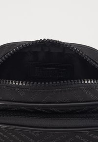 Svart dragkedjeväska med präglat logomönster, slät textur och en dragkedjefack med tyginsida.