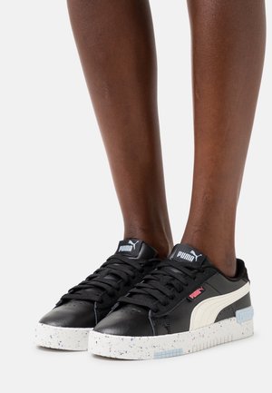 Jambes portant des baskets Puma noires avec une bande blanche et des semelles blanches mouchetées sur un fond clair uni.